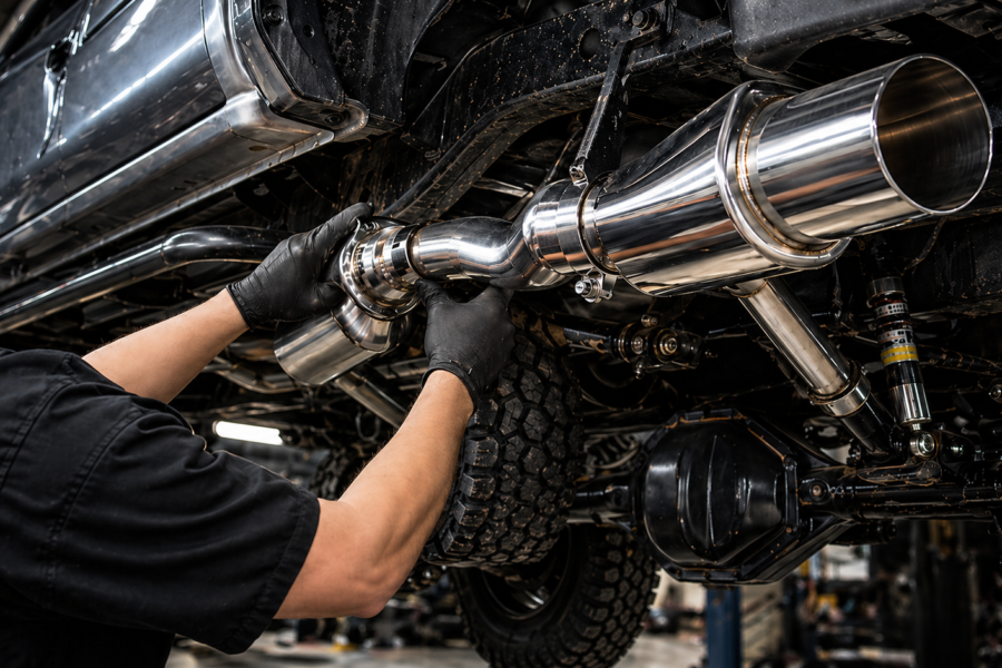 A mechanic wearing black gloves installs a high-quality, polished stainless steel heavy-duty diesel exhaust kit onto the undercarriage of a 4x4 diesel truck