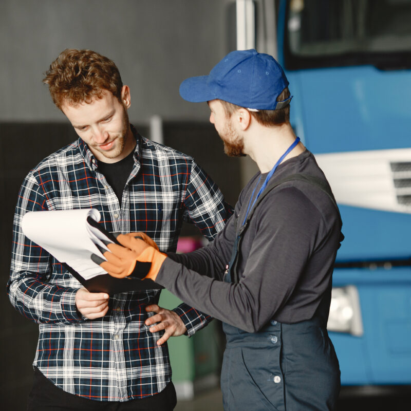 Two mechanics discussing the service checklist in front of a blue heavy-duty truck, emphasizing how often should a truck be serviced.